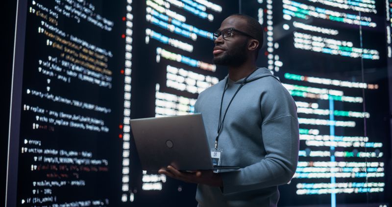 A man in a hoodie holding a laptop smiling with streams of code in the background